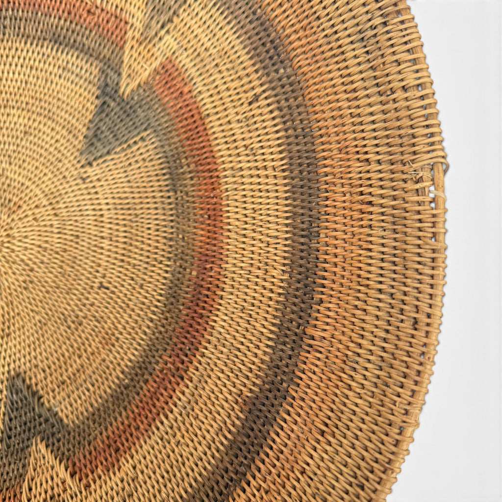 Close-up of a woven circular wall hanging or basket made from natural tan reed, with concentric geometric bands in brown, rust red, and dark charcoal on a white background.