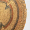 Close-up of a woven circular wall hanging or basket made from natural tan reed, with concentric geometric bands in brown, rust red, and dark charcoal on a white background.