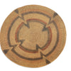 A round woven basket rug with a Southwestern geometric pattern in natural tan, brown, and reddish-brown on a white background.