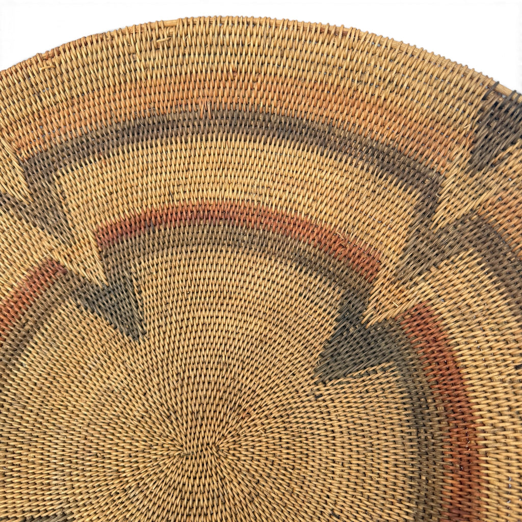 A close-up of a woven circular basket or tray made from natural tan reed fibers, featuring concentric geometric bands in dark brown, rust red, and charcoal gray.