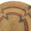 A close-up of a woven circular basket or tray made from natural tan reed fibers, featuring concentric geometric bands in dark brown, rust red, and charcoal gray.
