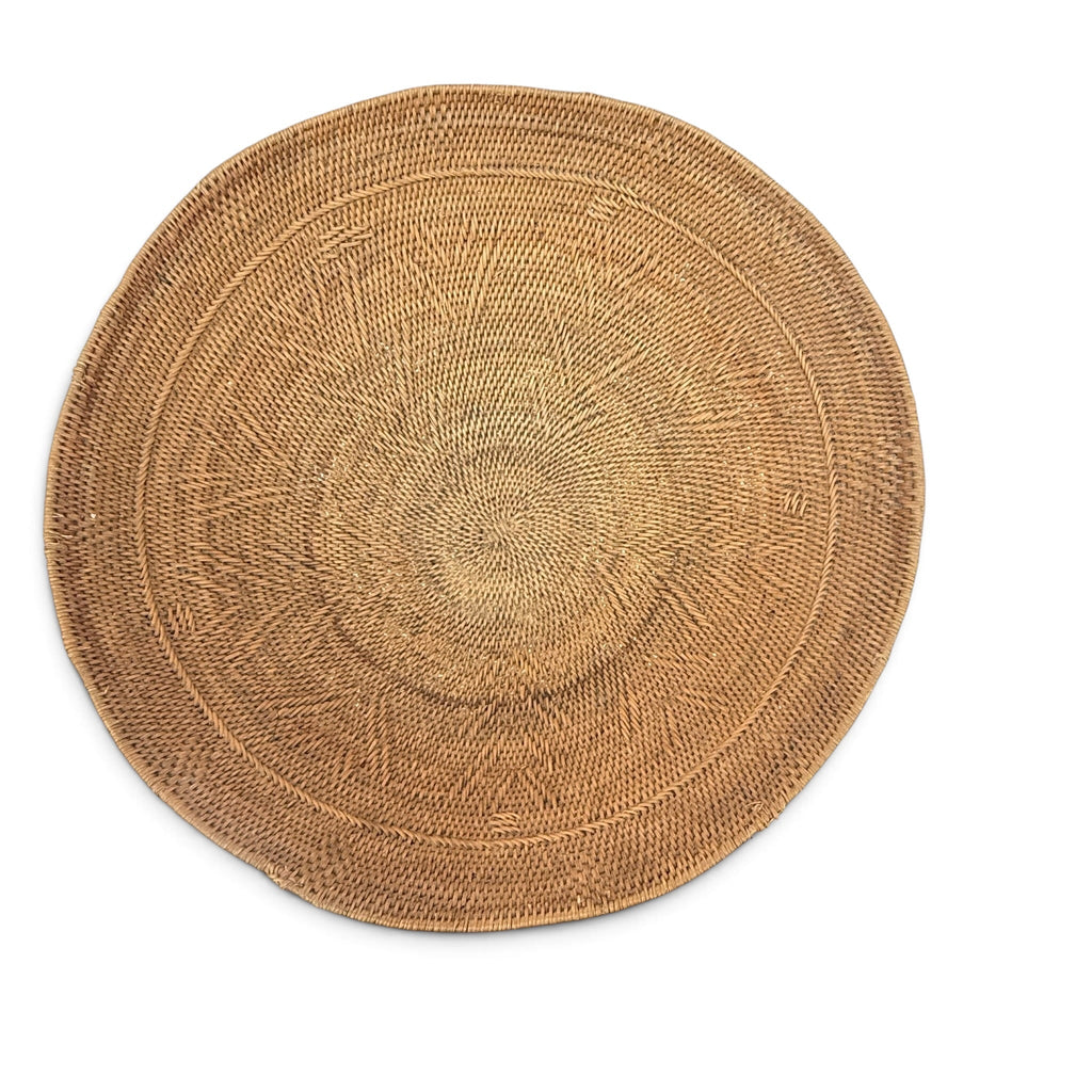 A round natural woven placemat or rug in a light tan color, featuring concentric circular patterns and a textured basket-weave design, shown on a solid white background.