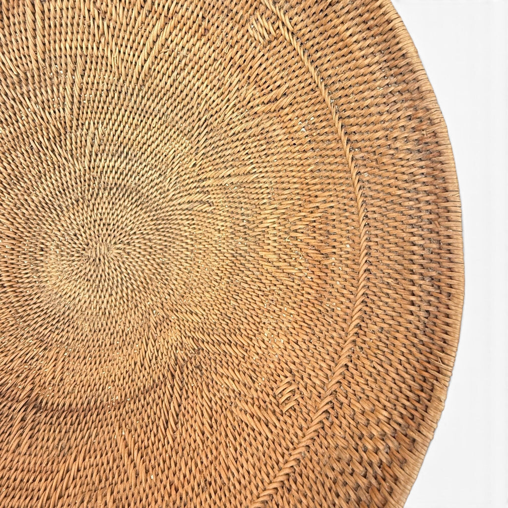 A close-up of a round woven natural fiber plate or tray, showing the detailed radial pattern of the wicker on a white background.